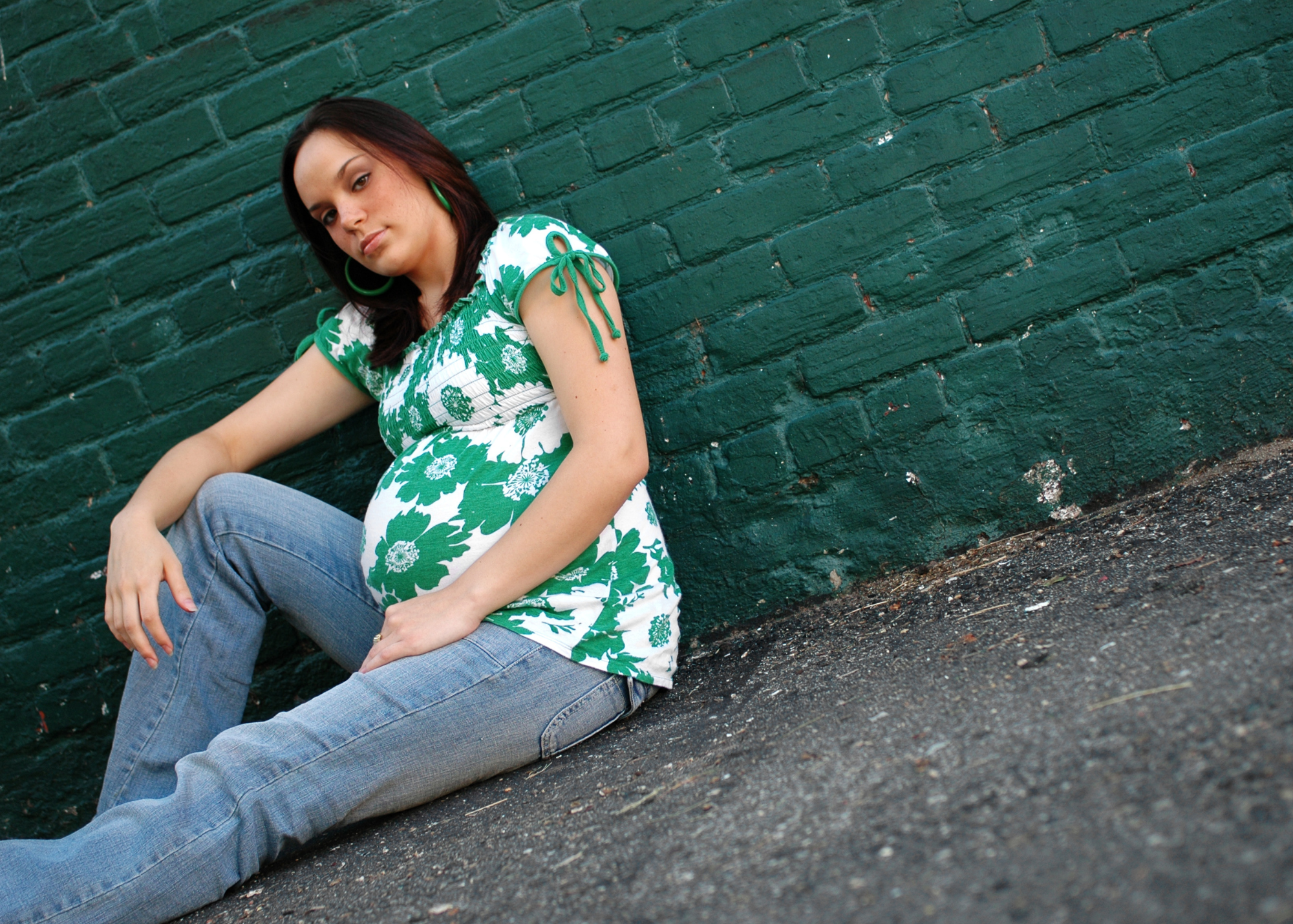 A pregnant person with a round belly leans against a green brick wall. They have shoulder length brown hair and are white. They are wearing a green and white floral top and light coloured jeans.